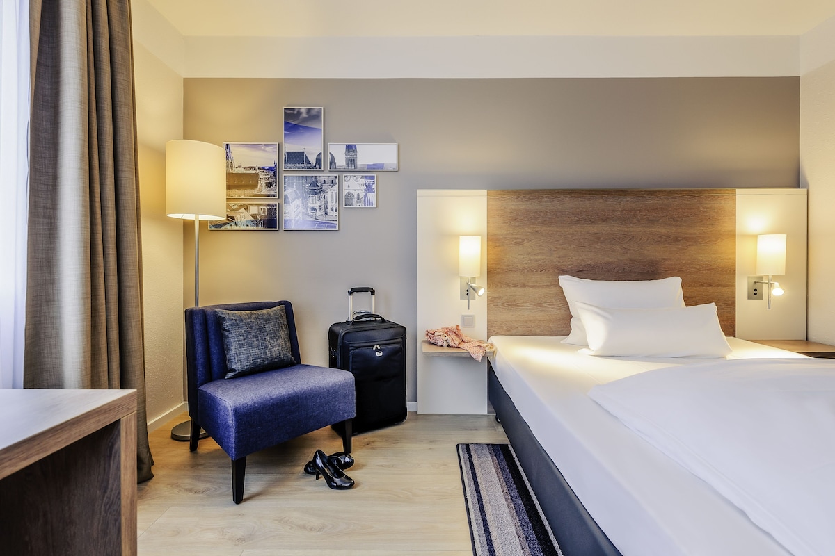 Una habitación de hotel con una cama, una silla, un escritorio, una maleta y decoración en la pared.
