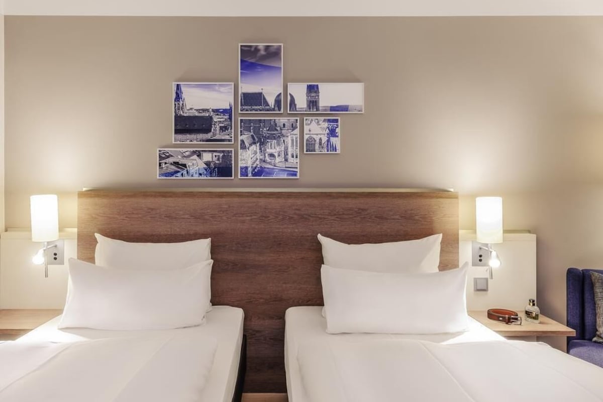 Habitación de hotel con dos camas, cabecero de madera, obra de arte colgada en la pared, lámparas de noche y una pequeña mesa con lámpara.