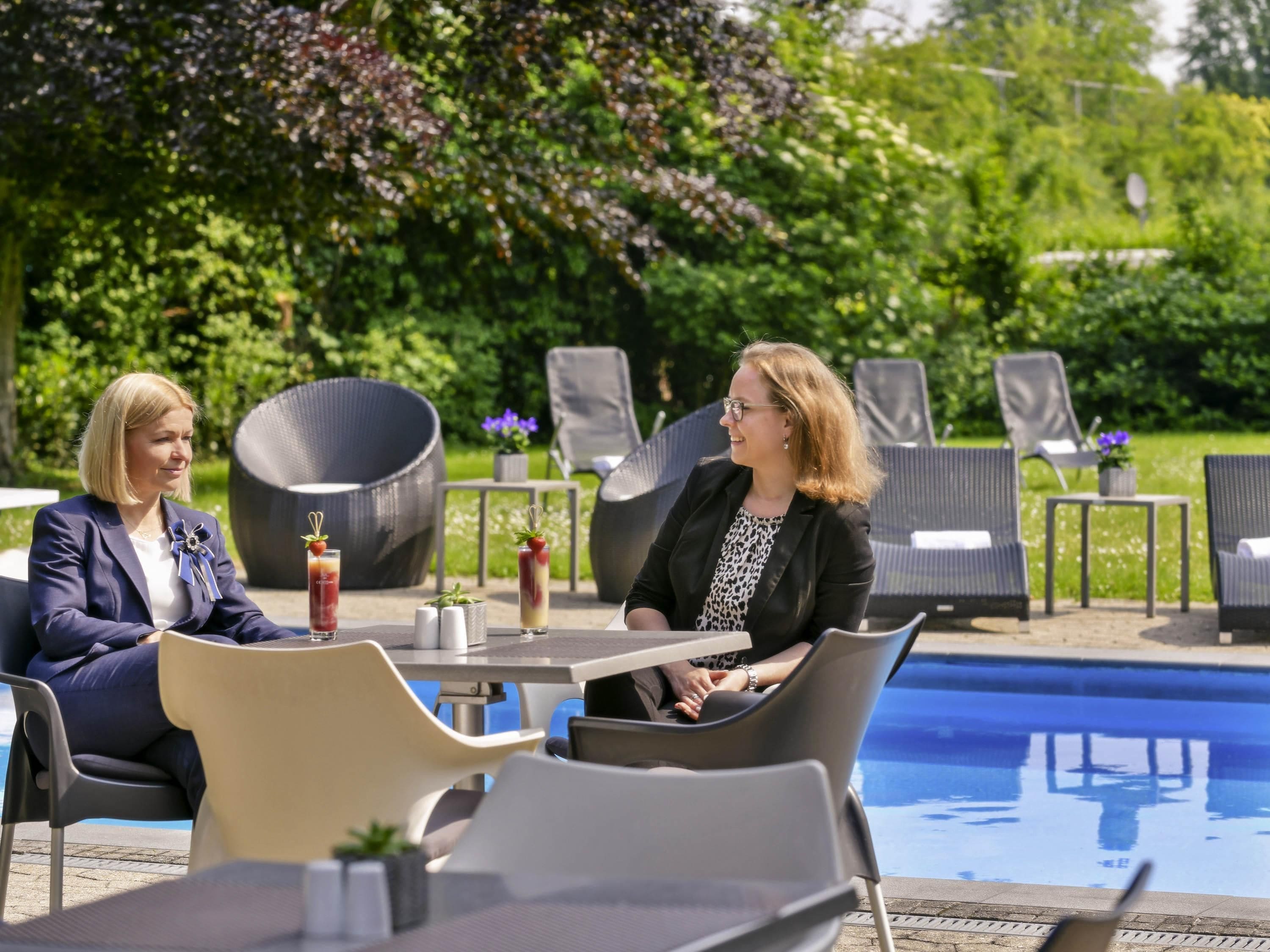 Two people seated at a table by a poolside with lounge chairs and a garden in the background.