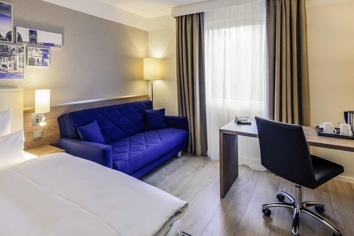 Quarto de hotel com cama, sofá azul, escrivaninha com cadeira, abajur e telefone na escrivaninha.