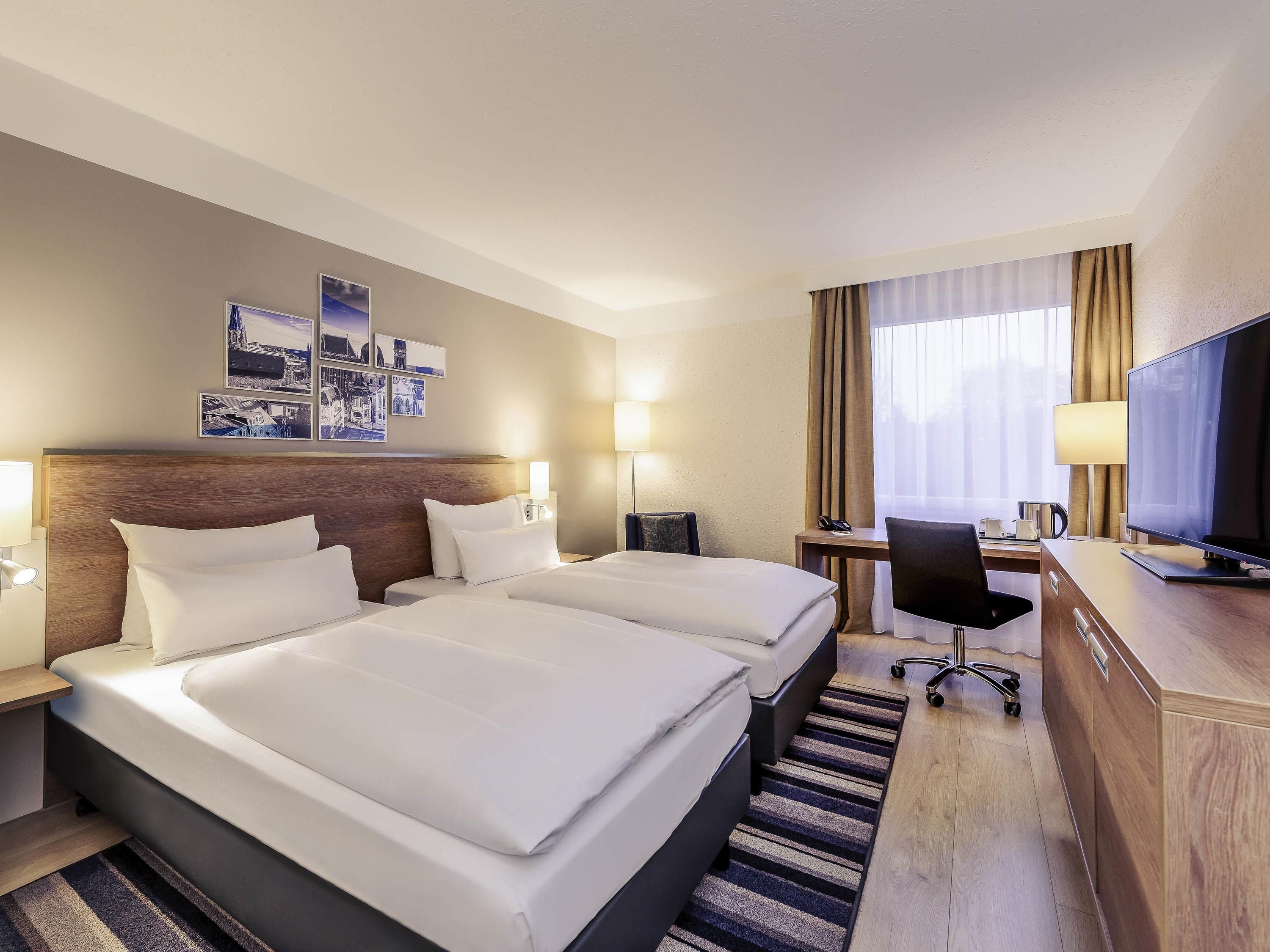 Habitación de hotel con dos camas, un escritorio con silla, televisión y ventana con cortinas.