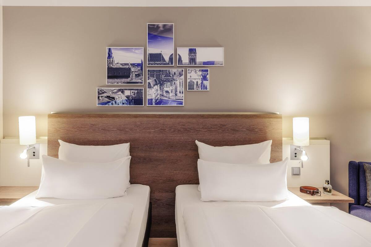 Quarto de hotel com duas camas, cabeceira de madeira, obras de arte na parede e abajures nas mesas de cabeceira.