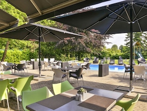 An outdoor seating area with tables and chairs, umbrellas, and a pool in the background. - Mercure Hotel Aachen Europaplatz (Aachen)