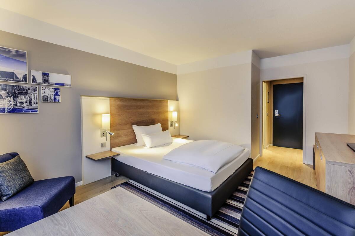 Quarto de hotel com cama, mesas de cabeceira, escrivaninha, cadeira e porta.