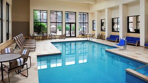 An indoor swimming pool with lounge chairs and tables, large windows, and a view of the outside.