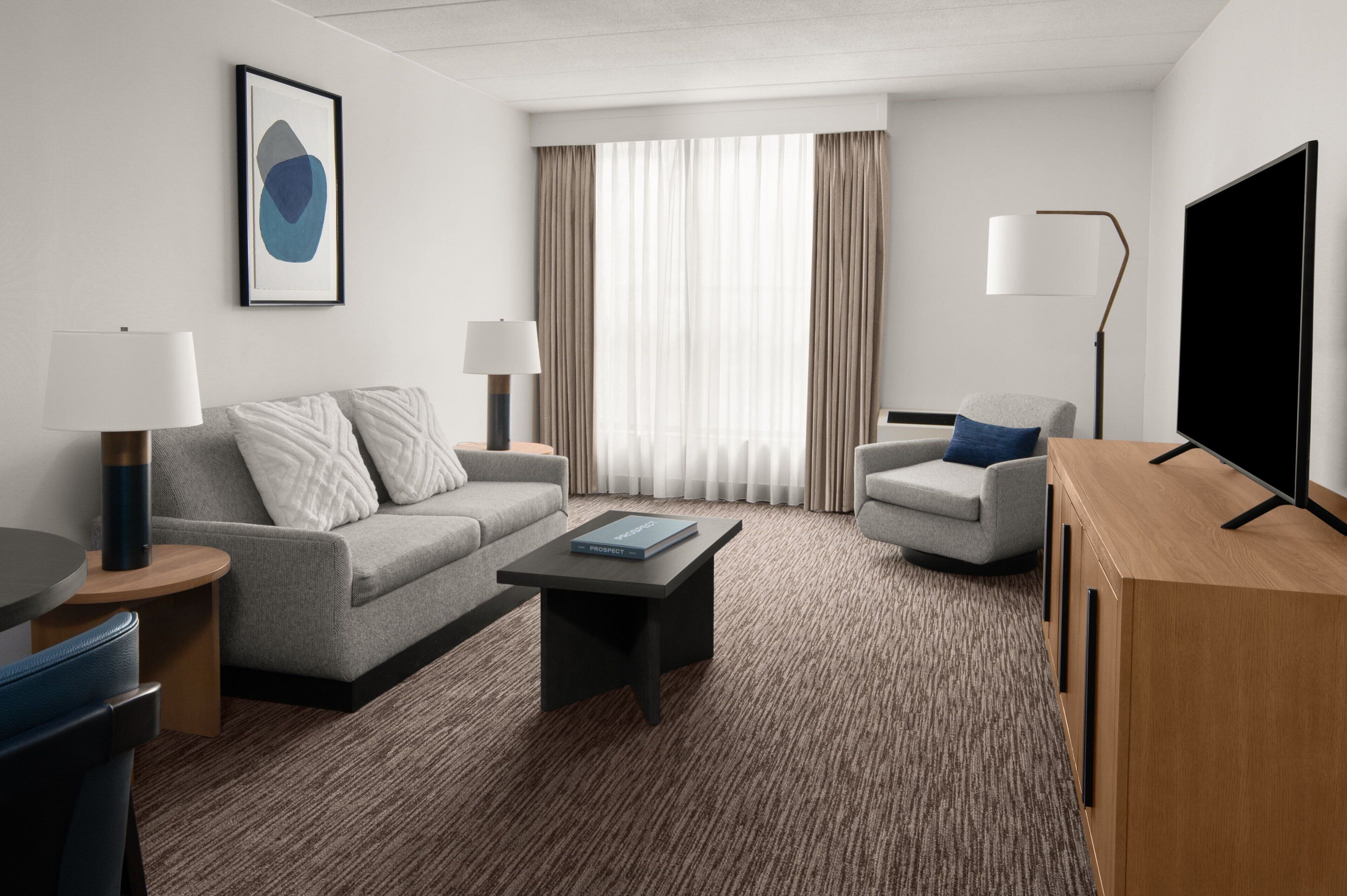 A modern hotel room with a grey sofa, a small table, a coffee table, a TV, and a lamp.