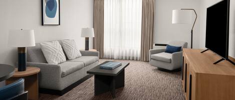 Une chambre d’hôtel moderne avec un canapé gris, une petite table, une table basse, un téléviseur et une lampe.