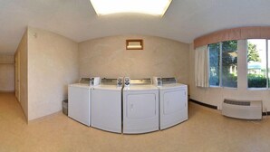 Ein Waschraum mit vier gestapelten Waschmaschinen und Trocknern, einem kleinen Fenster mit Vorhängen und einer Lüftung an der Wand.