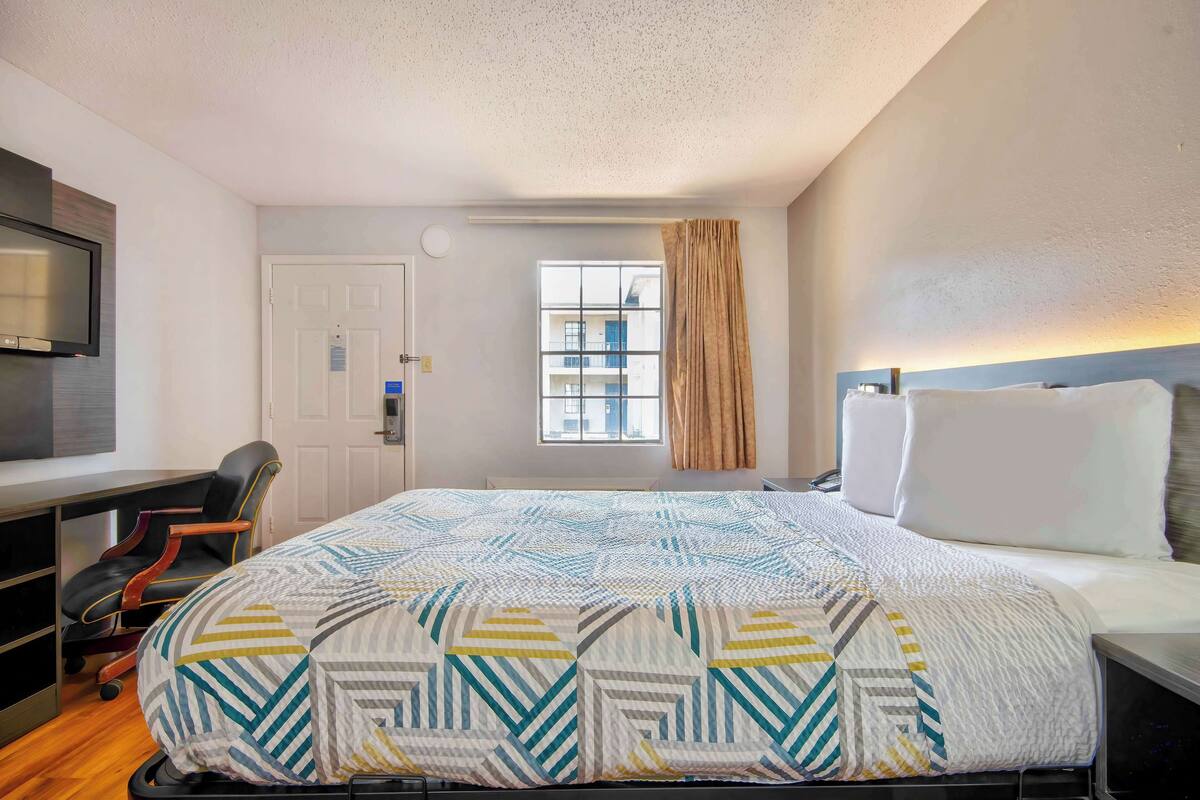 A hotel room with a bed, a desk, a chair, a TV, a window with curtains, and a view of buildings outside.