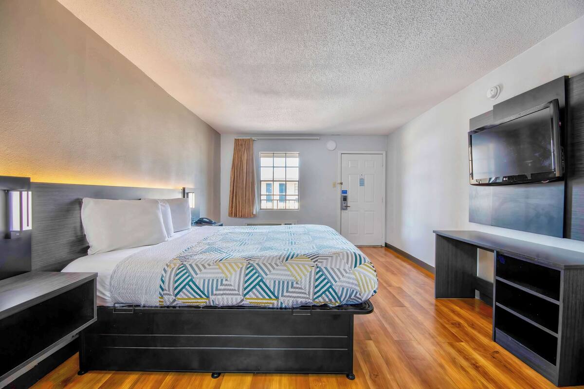 A modern hotel room with a bed, bedside table, TV, and a window with curtains.