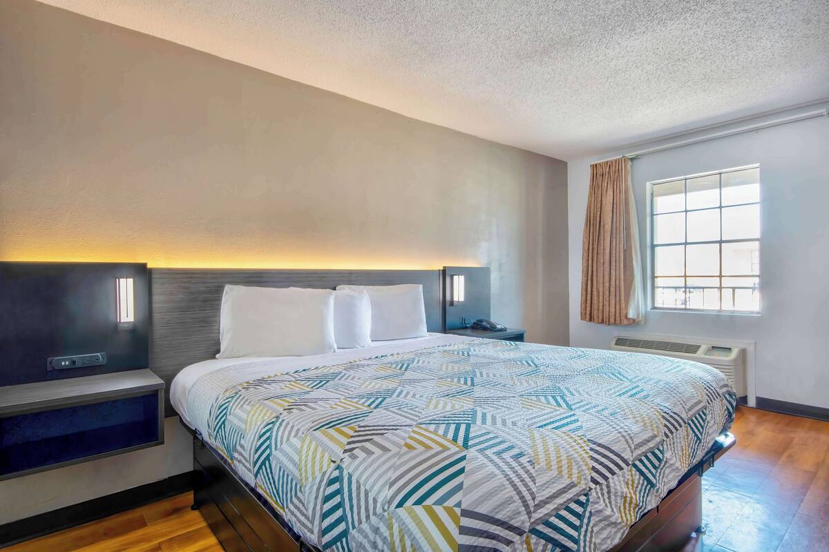 A modern hotel room with a large bed, bedside tables, a built-in heater, a window with curtains, and a view of the outdoors.