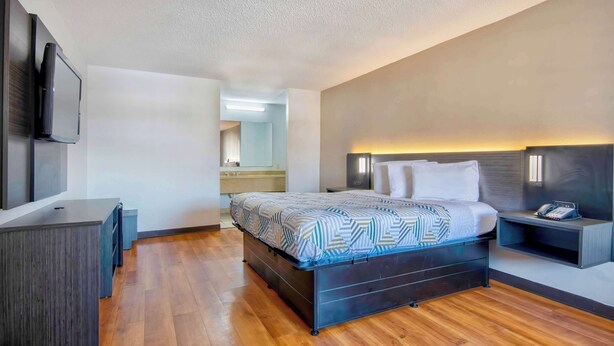 A modern hotel room with a wooden floor, a bed with patterned bedding, a bedside table with a phone, and a TV mounted on the wall.