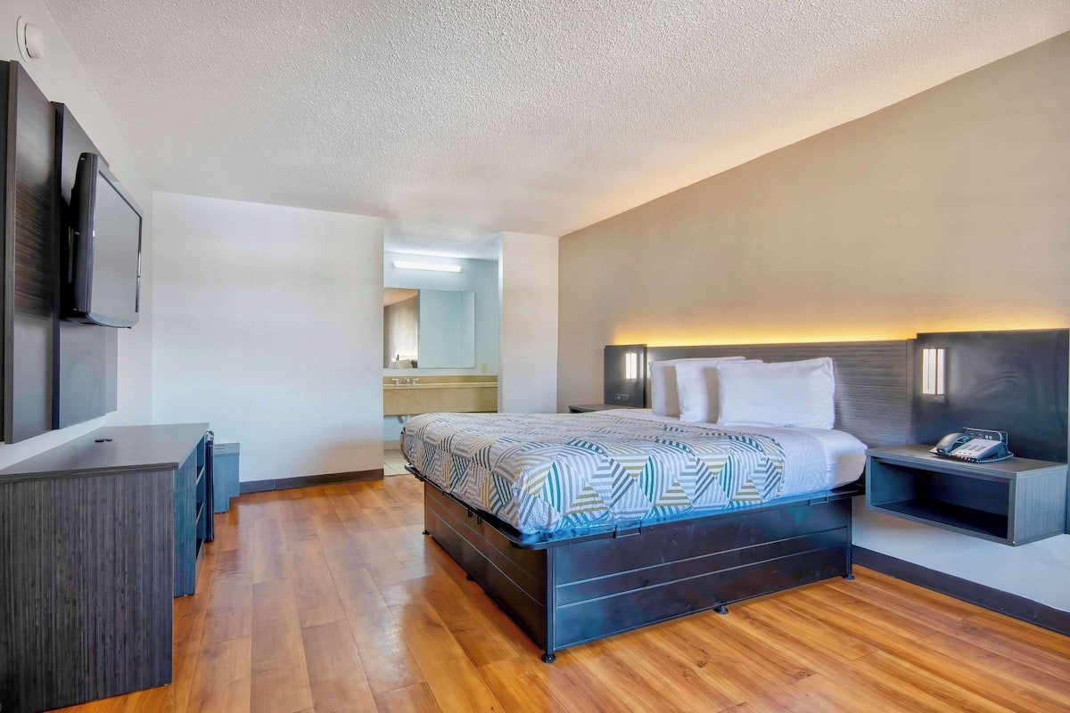 A modern hotel room with a wooden floor, a bed with patterned bedding, a bedside table with a phone, and a TV mounted on the wall.