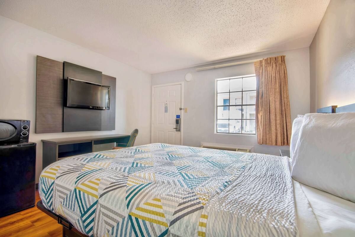 A modern hotel room with a bed, a desk, a flat-screen TV, and a window with curtains.