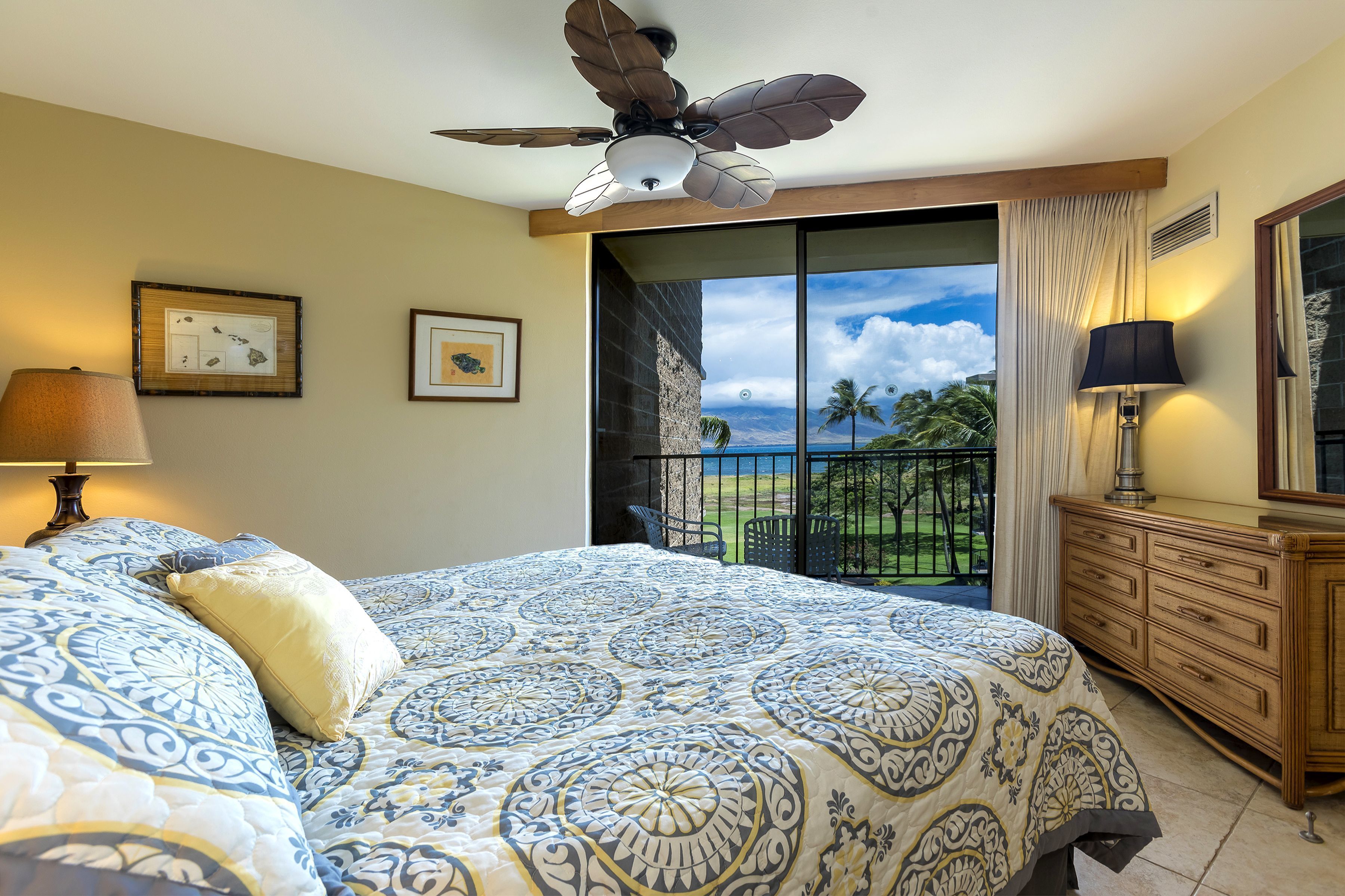 A bedroom with a large bed, a ceiling fan, a balcony with a view of palm trees and a grassy area, and a wooden dresser.