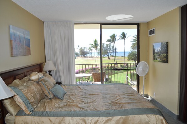 A hotel room with a large bed, a balcony with a view of the ocean and palm trees, and a fan. - Kauhale Makai - Maui Condo & Home (Kihei)
