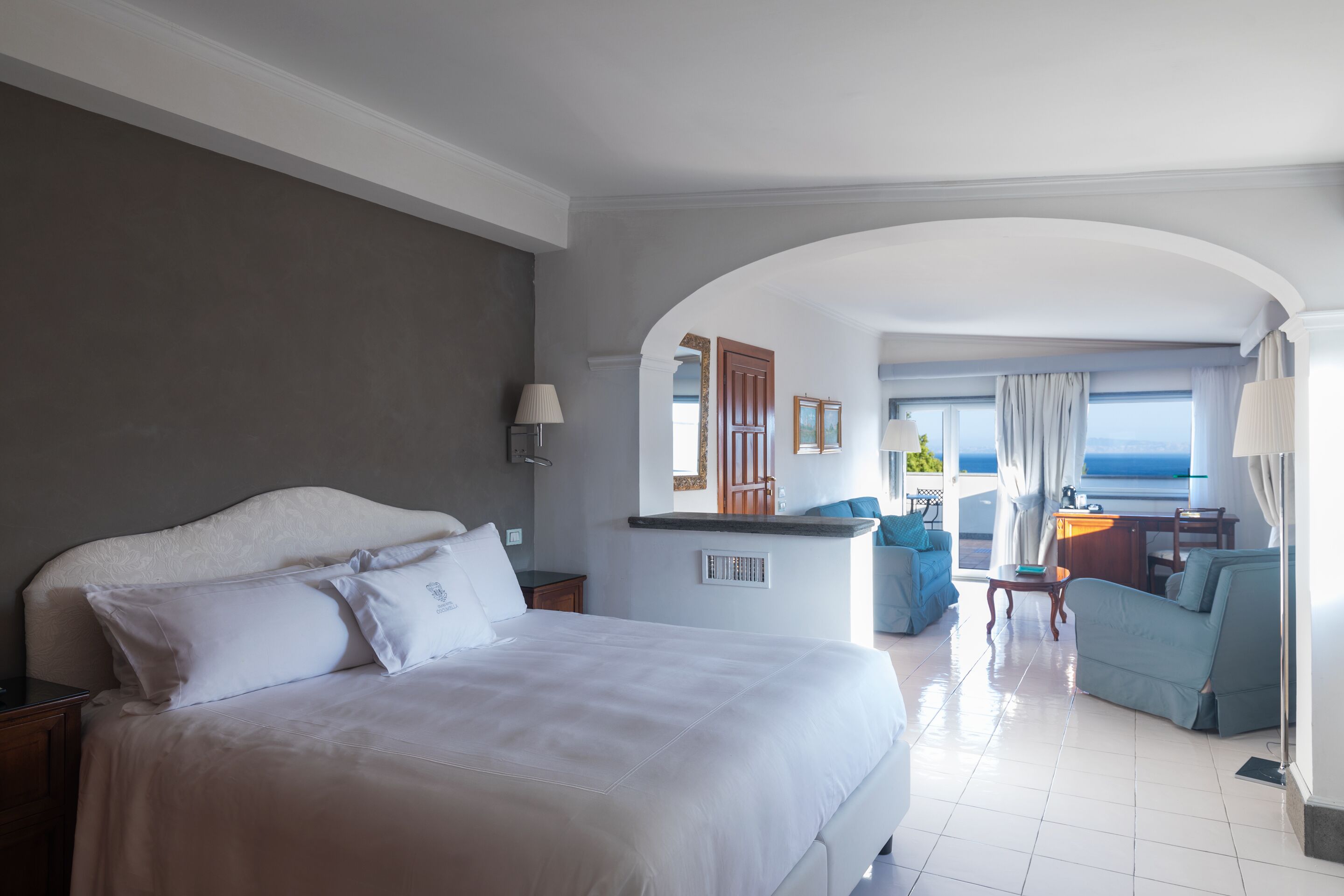 A hotel room with a large bed, a desk, a chair, and a view of the sea.
