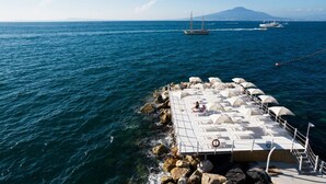 A pier with a deck area featuring lounge chairs and umbrellas, overlooking a body of water with boats and a distant mountain. - Grand Hotel Cocumella (Sant'Agnello)