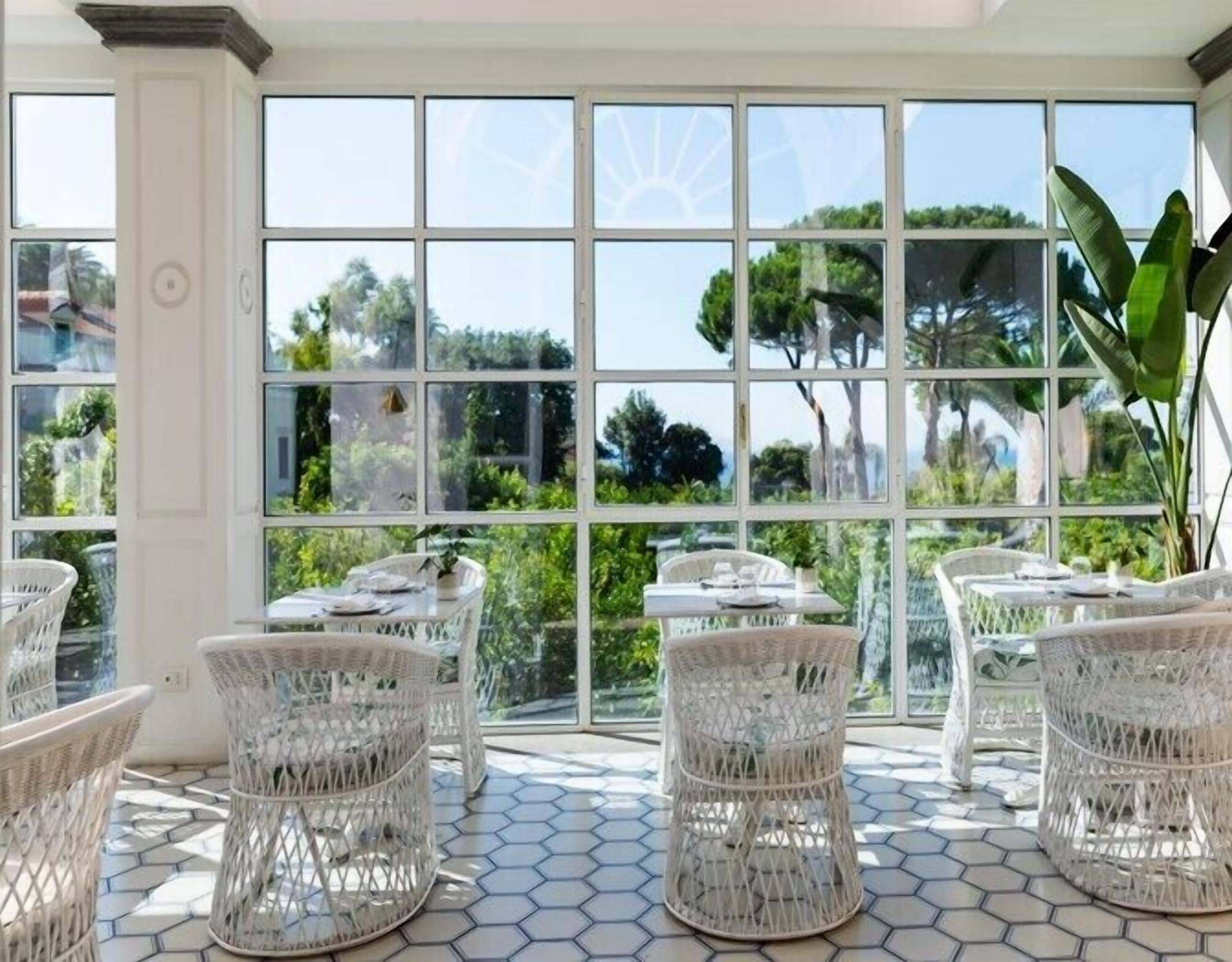 A sunlit patio with hexagonal tiled flooring, white wicker furniture, and large windows offering a view of greenery and trees.