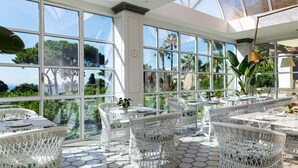 A sunlit patio with white wicker furniture and large windows offering a view of palm trees and the sea. - Grand Hotel Cocumella (Sant'Agnello)