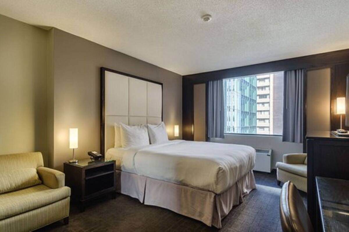 A hotel room with a large bed, a bedside table with a lamp, a chair, and a view of the city through the window.