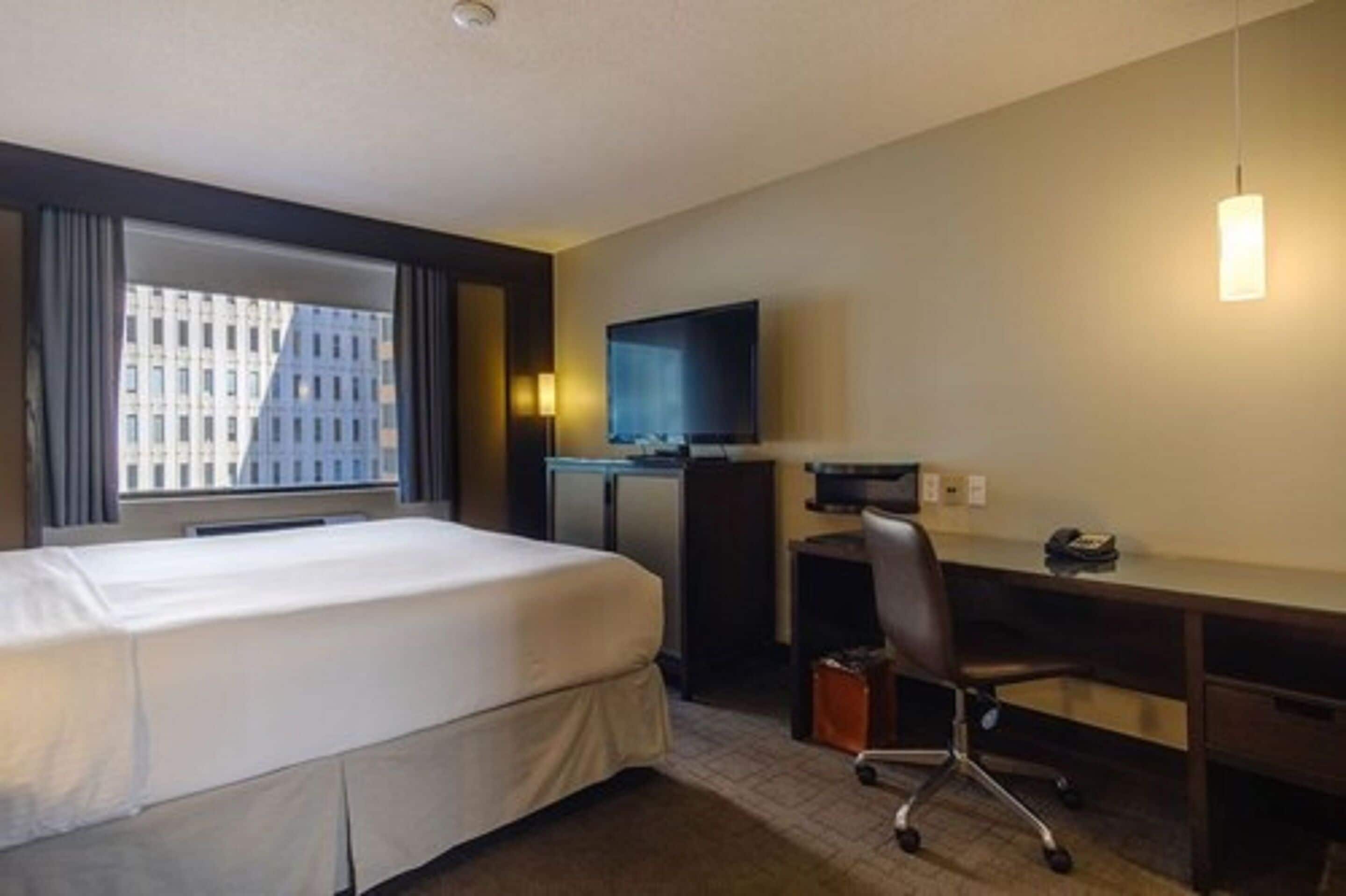 A hotel room with a bed, a desk, a chair, a TV, and a window with a city view.