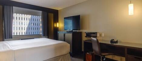 A hotel room with a bed, a desk, a chair, a TV, and a window with a city view.