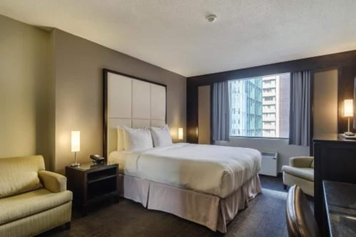 A hotel room with a large bed, a bedside table, a chair, a desk, and a window with curtains.