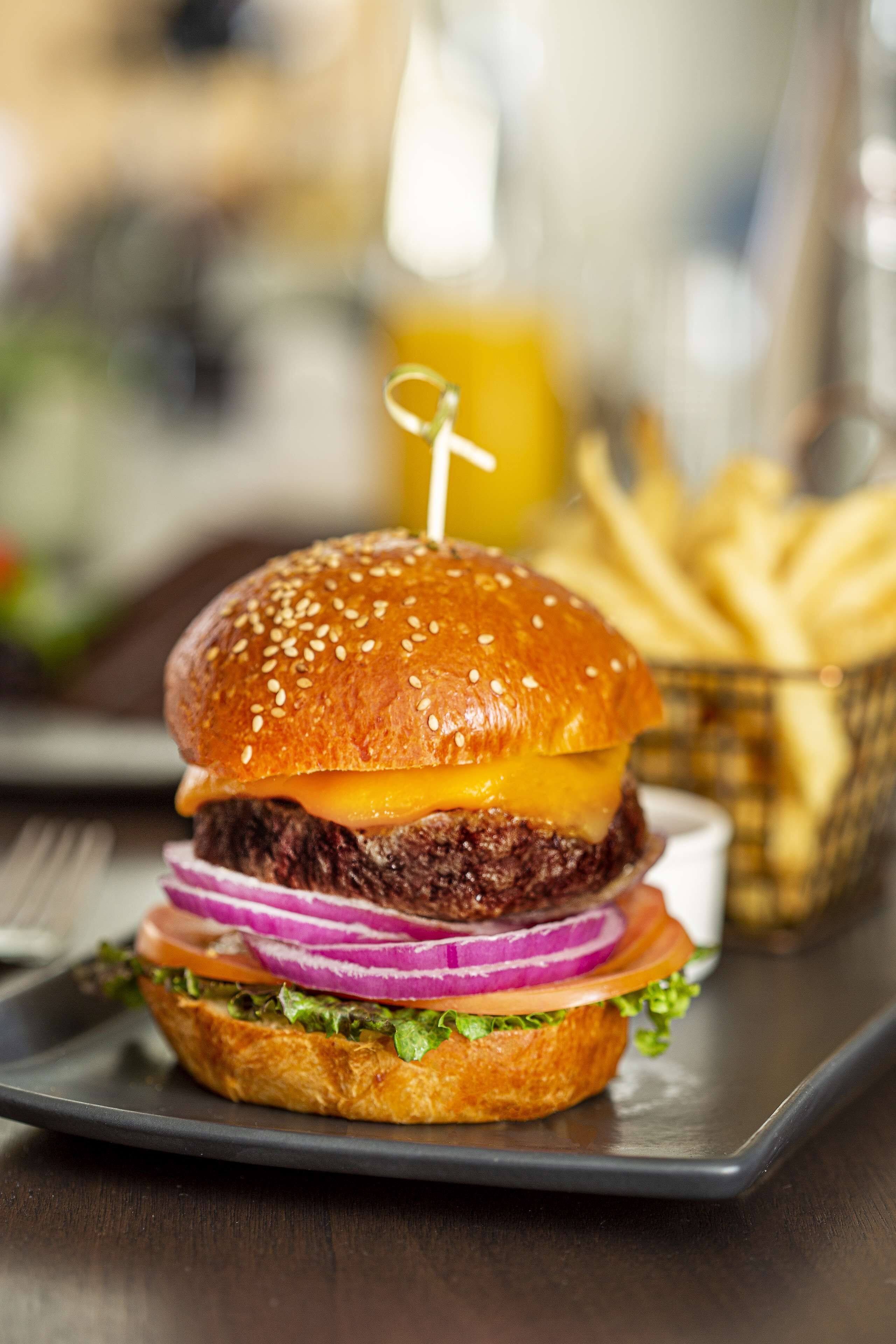A burger with a sesame seed bun, cheese, lettuce, tomato, and red onion, served on a dark plate.