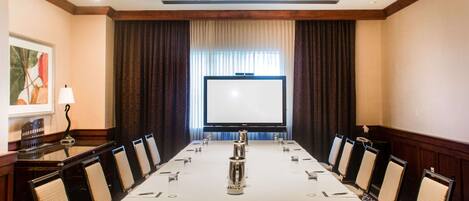 A conference room with a long table, chairs, a projector screen, and a large window with curtains.