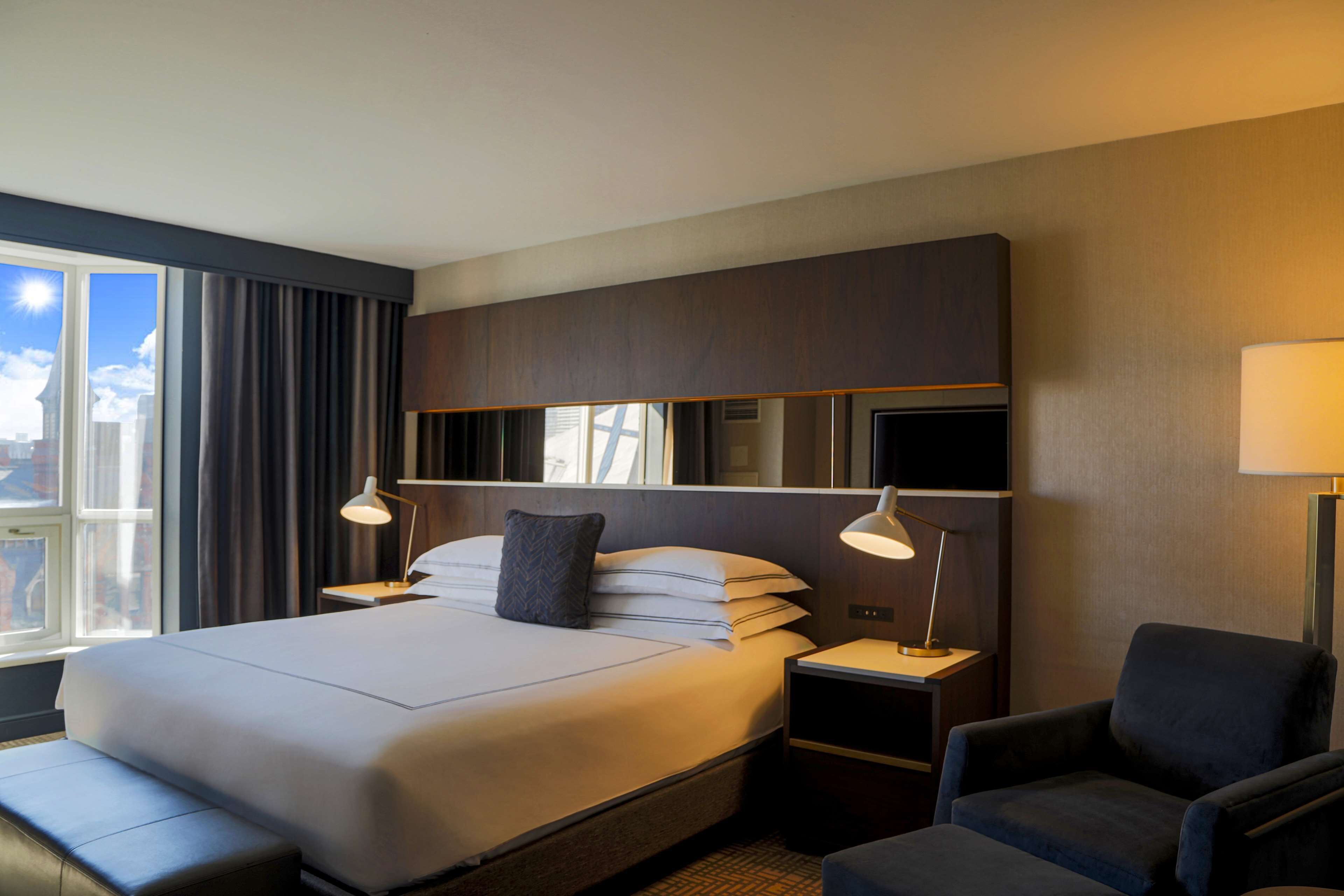 Habitación de hotel con una cama grande, mesitas de noche con lámparas, una silla y vistas de la ciudad a través de la ventana.