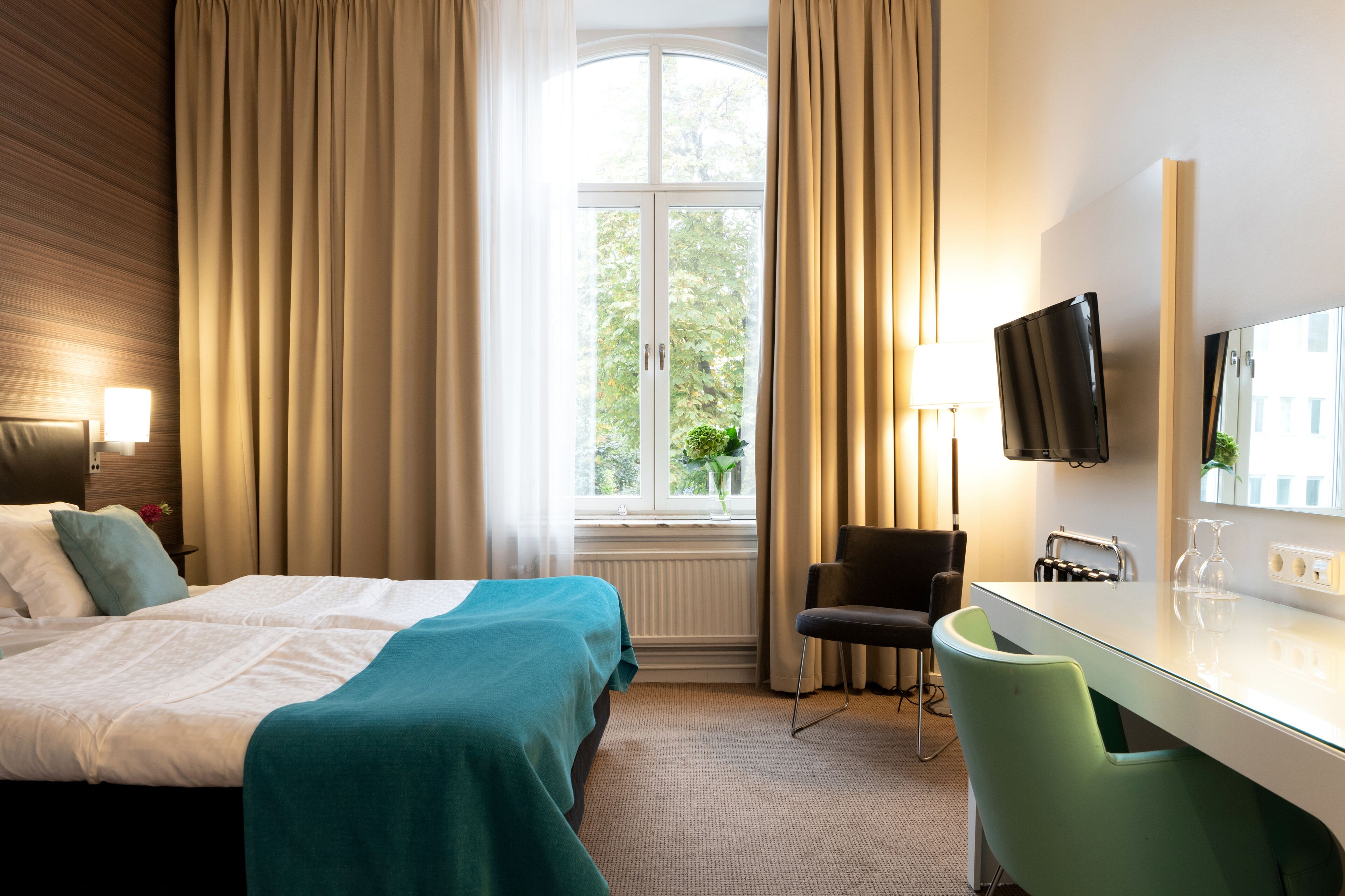 A hotel room with a bed, a desk, a chair, a TV, and a window with curtains.