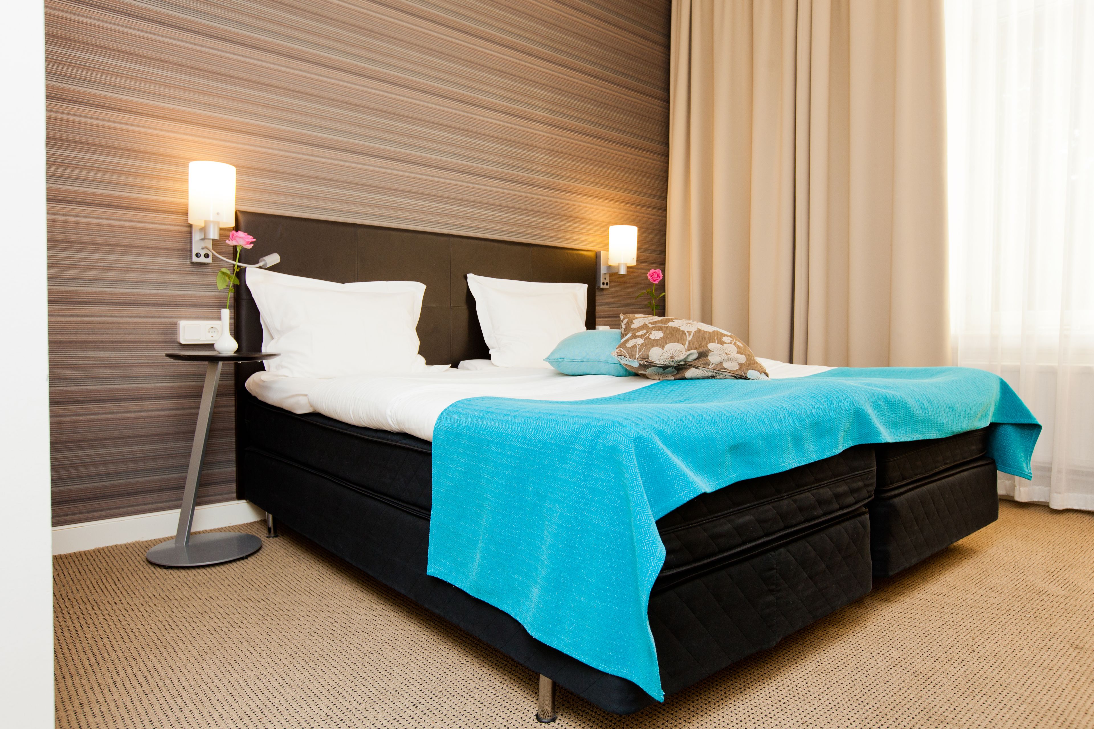 Habitación de hotel con una cama, dos lámparas de noche, una mesita y un jarrón con flores.
