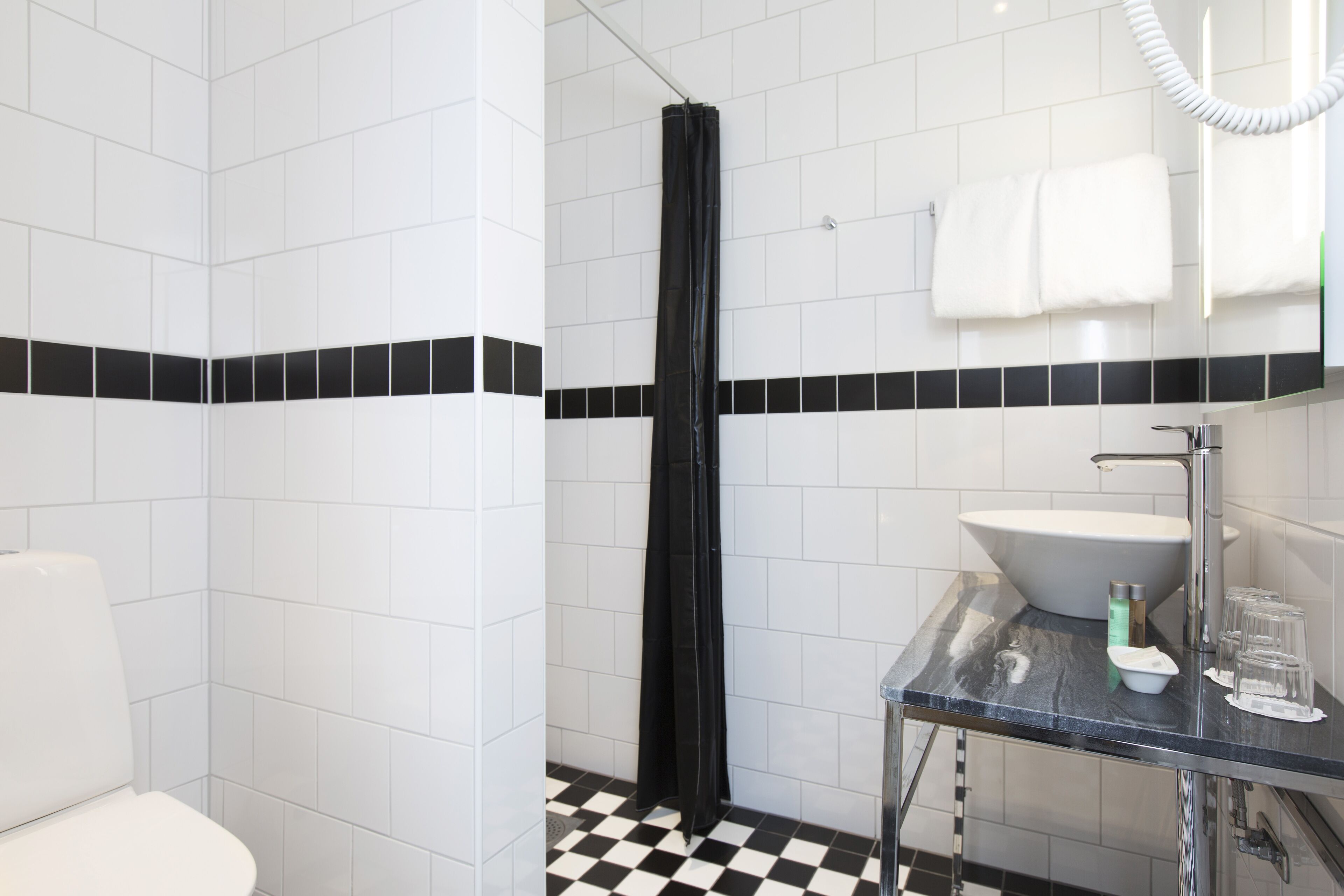 Une salle de bain avec des carreaux blancs, un sol à carreaux noirs et blancs, un rideau de douche noir, un lavabo blanc et un miroir sur lequel est accrochée une serviette.