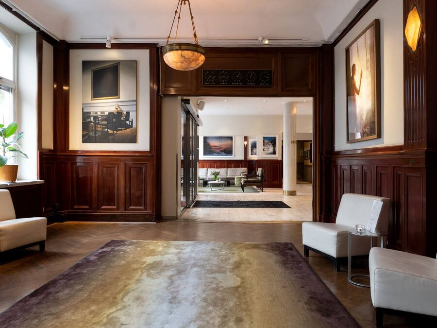 A spacious lobby with a large rug, wooden paneling, and a chandelier. There are several seating areas and artwork on the walls.