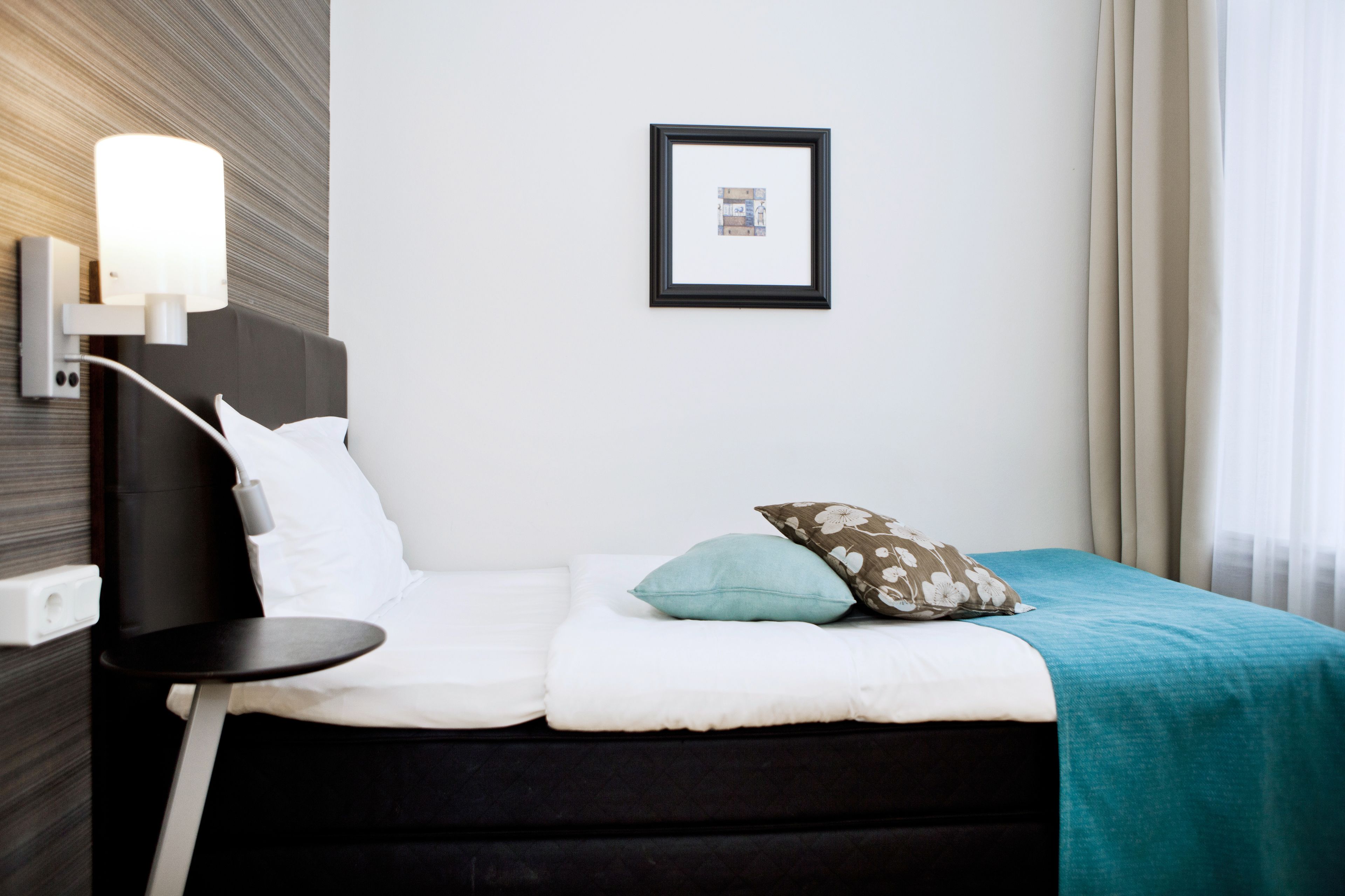 A hotel room with a bed, a bedside table, a wall-mounted lamp, and a framed picture on the wall.