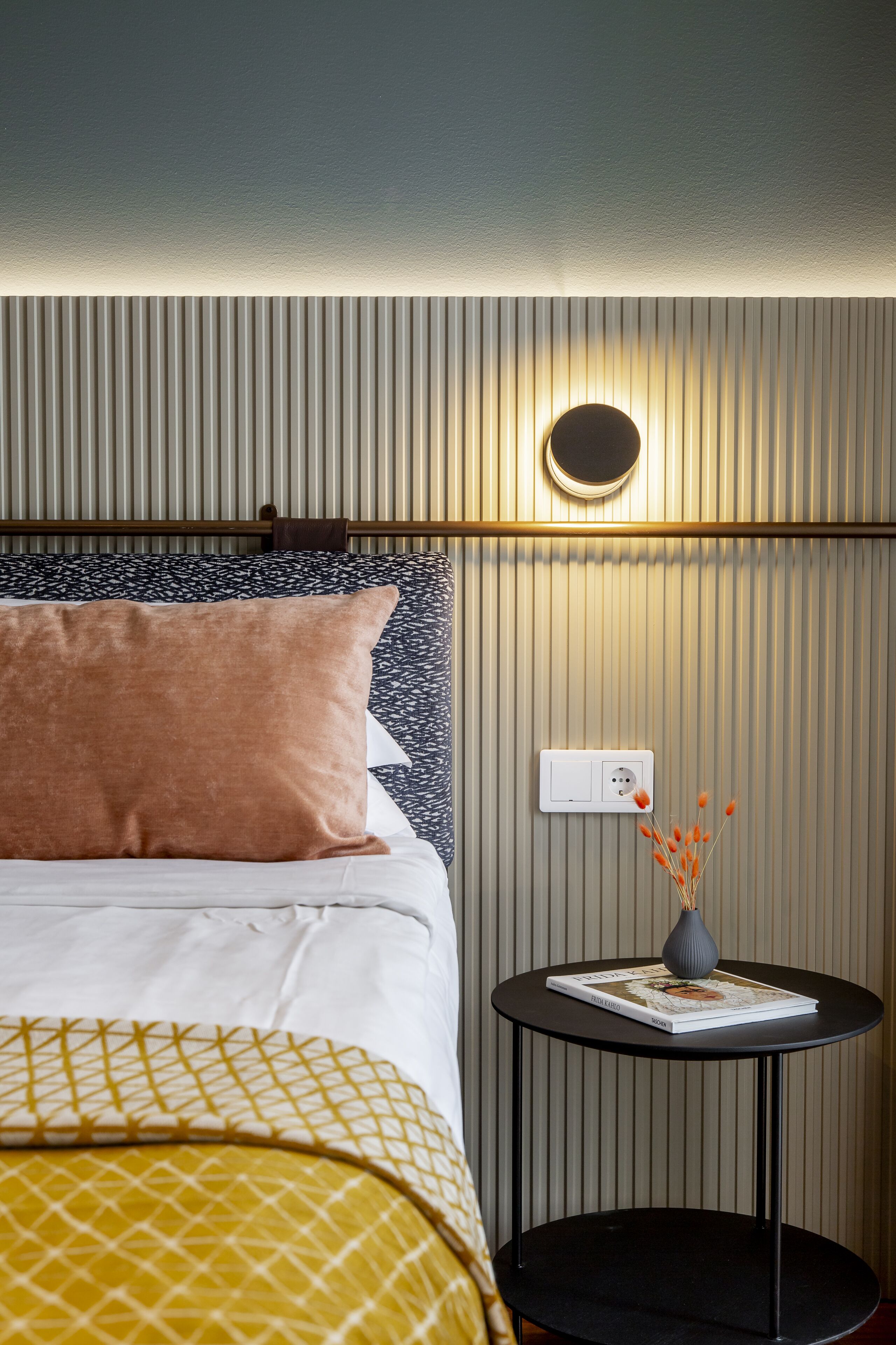 A neatly made bed with patterned bedding, a bedside table with a book and a vase, and a wall-mounted light fixture.