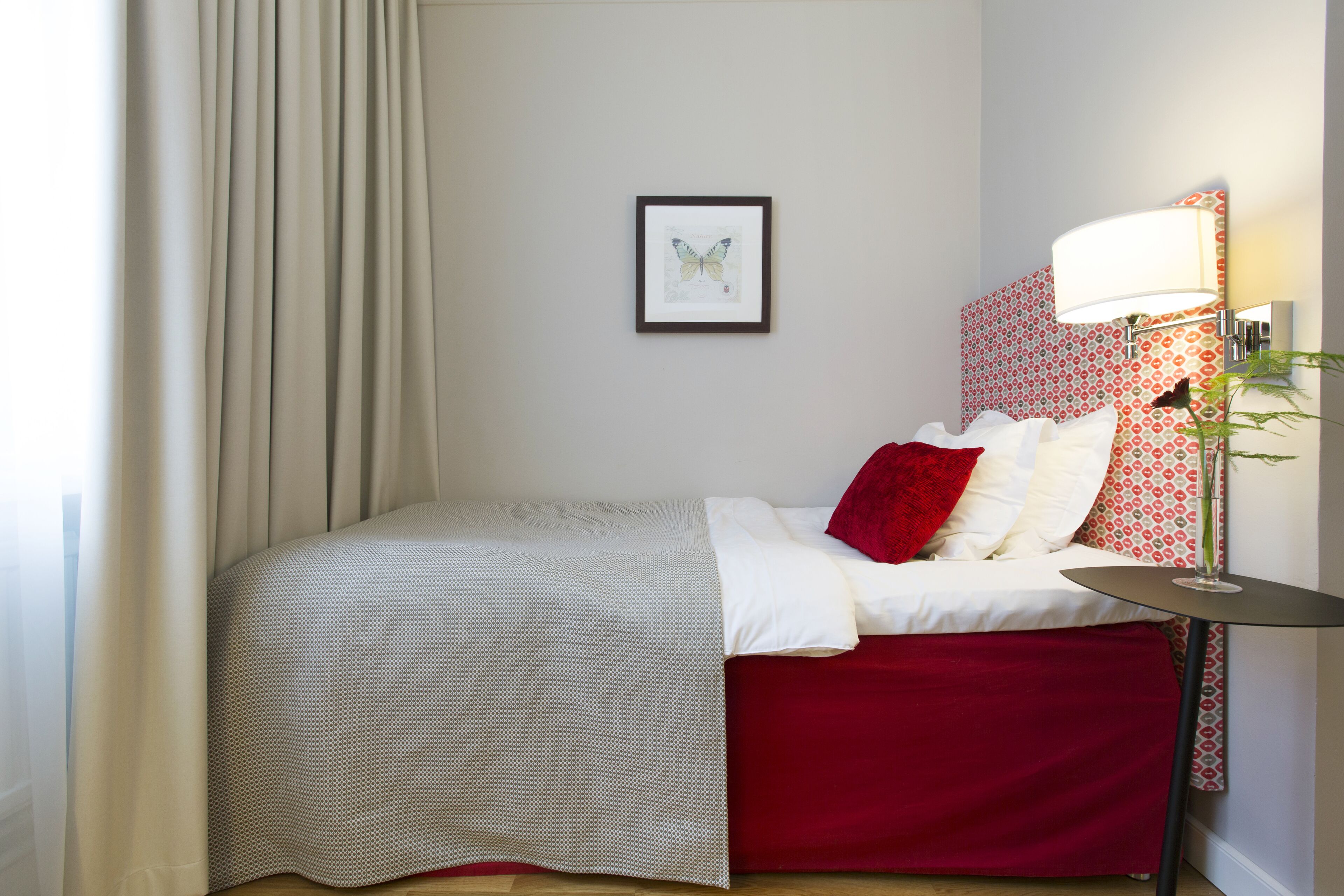 Una cama bien hecha con colcha roja y blanca, una obra de arte de una mariposa enmarcada en la pared y una lámpara con pantalla estampada.