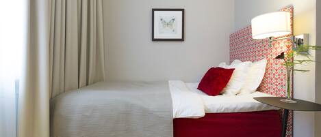 Una cama bien hecha con colcha roja y blanca, una obra de arte de una mariposa enmarcada en la pared y una lámpara con pantalla estampada.