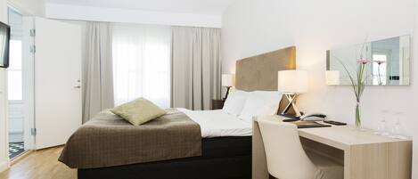 A hotel room with a bed, a desk, a chair, a lamp, a mirror, and a vase with flowers.