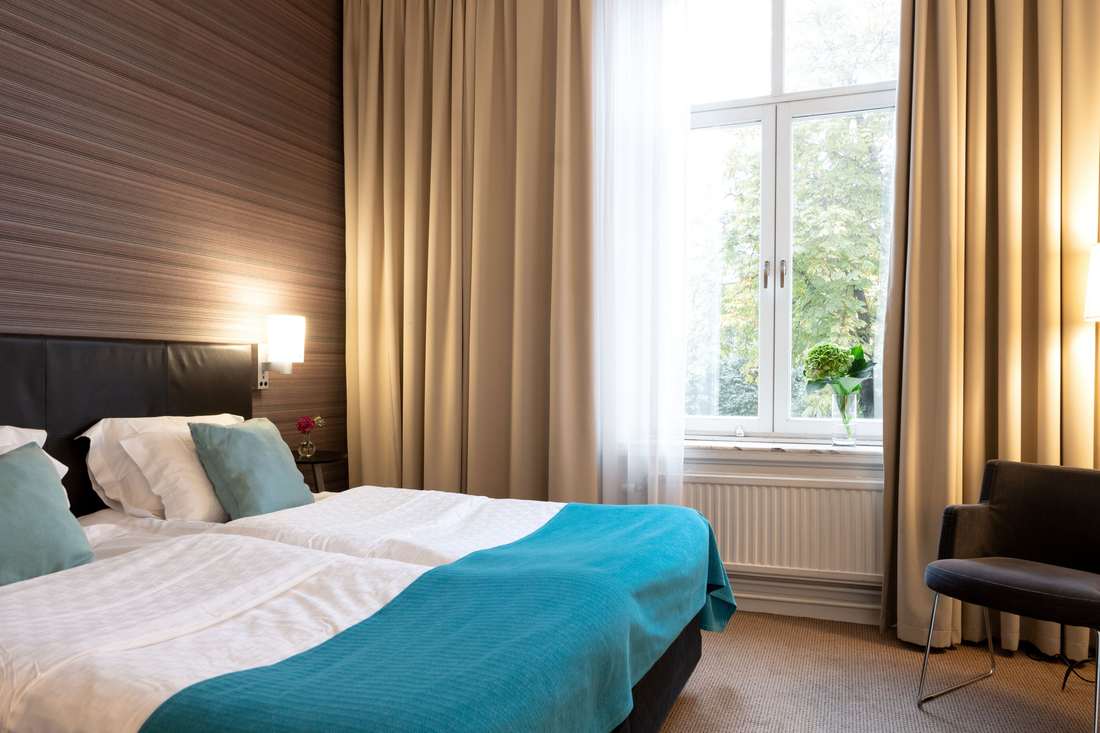 A hotel room with a large bed, two bedside tables, a chair, a window with curtains, and a view of greenery outside.