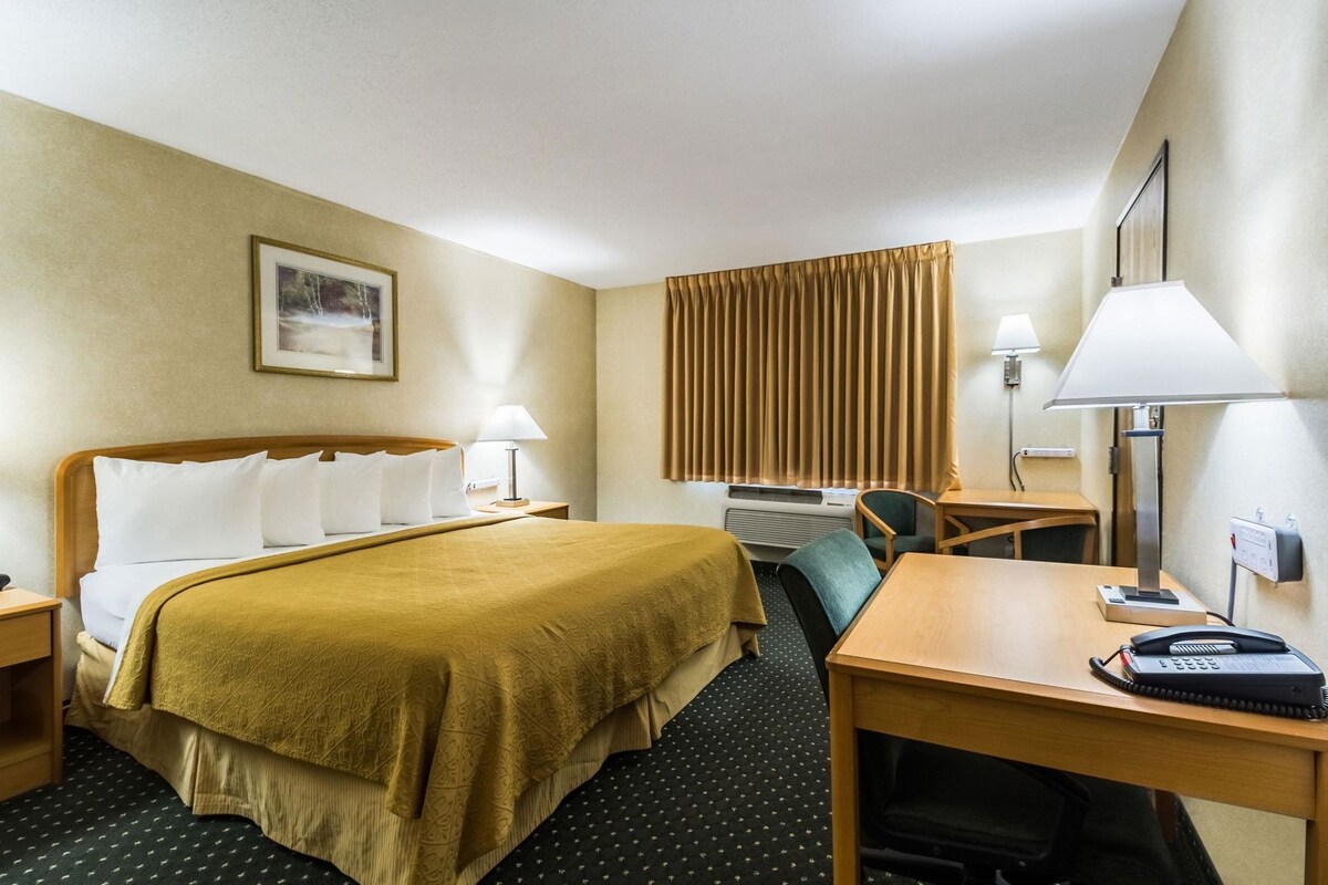 A hotel room with a bed, bedside tables, a desk, a chair, a lamp, a phone, and a window with blinds.