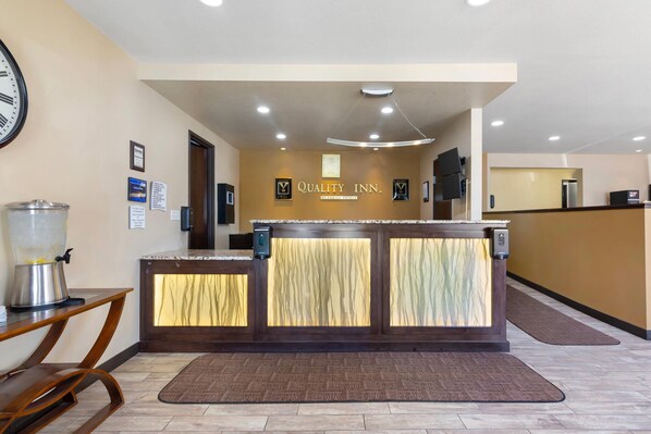 A reception desk with a sign reading "Quality Inn." There is a clock on the wall, a coffee machine, and a trash can.