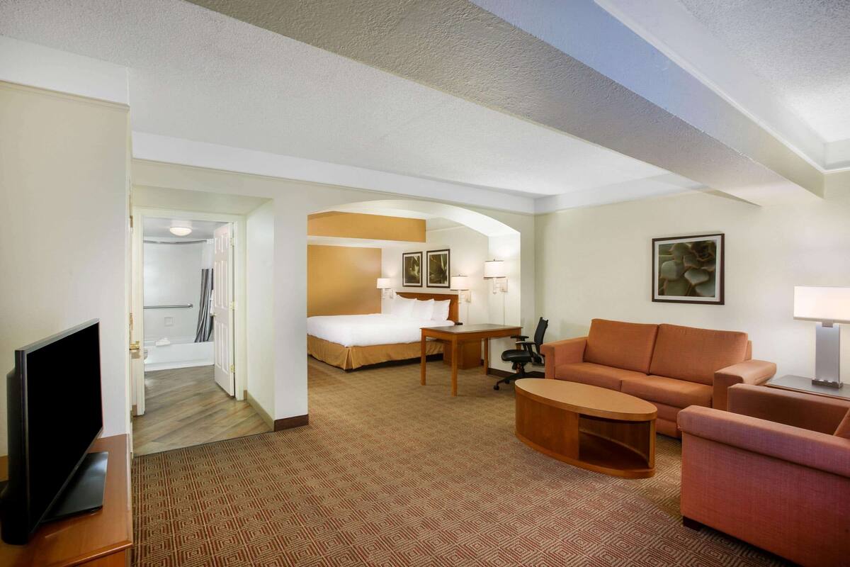 Habitación de hotel con cama, escritorio, sofá, televisión y baño visible a través de una puerta abierta.