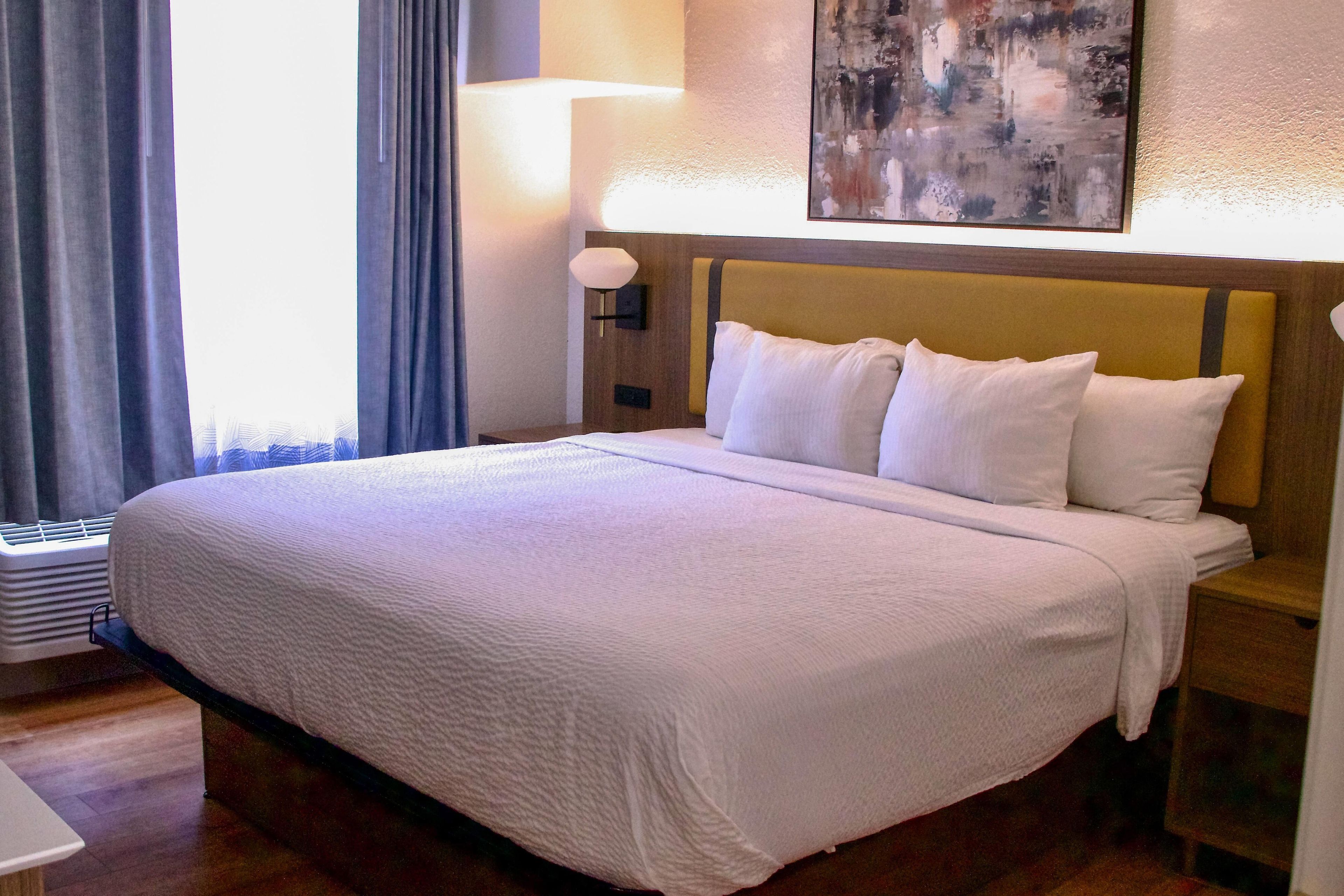 A hotel room with a bed, a nightstand, a lamp, a painting on the wall, and a window with curtains.