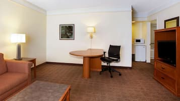 A hotel room with a sofa, a small table, a TV stand, a desk with a chair, a lamp, a microwave, and a refrigerator.