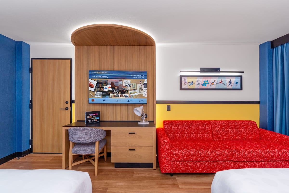 Ein Hotelzimmer mit Holzschreibtisch, einem roten Sofa, einem Bett und einem an der Wand befestigten Fernseher, der einen Reiskanal zeigt.