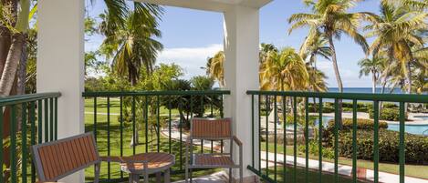 A balcony with two wooden chairs and a table, overlooking a pool and palm trees.