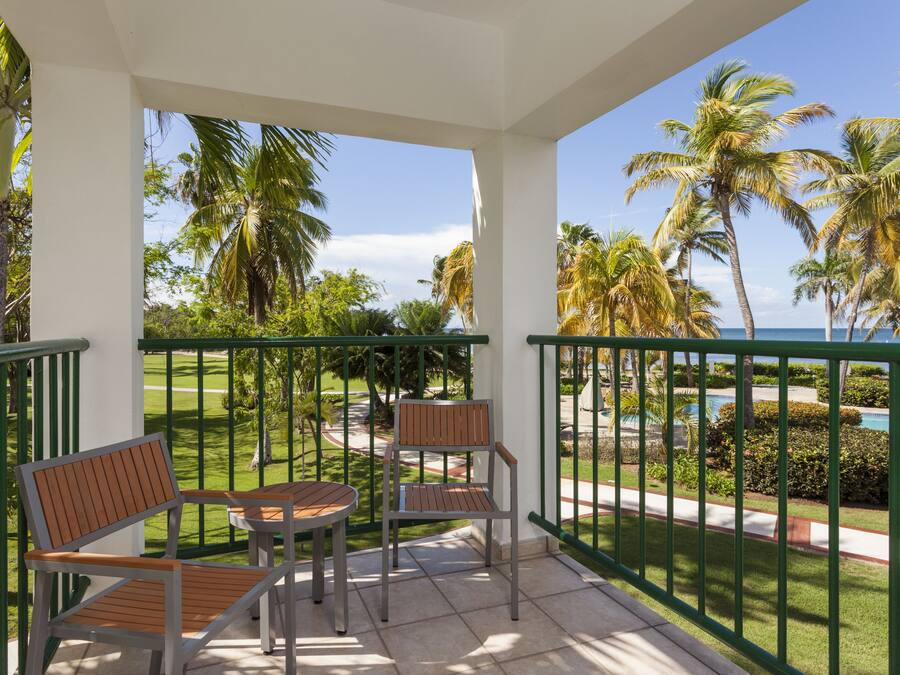 A balcony with two wooden chairs and a table, overlooking a pool and palm trees.