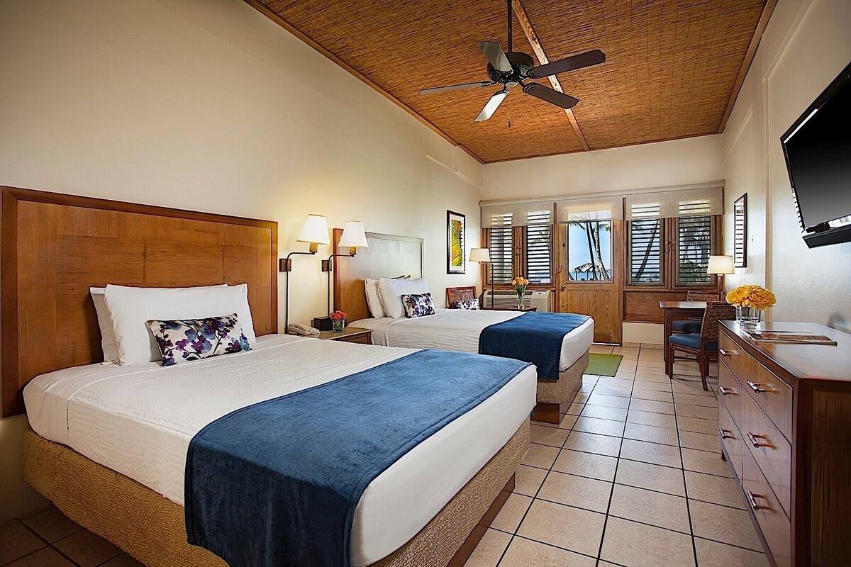 Ein Hotelzimmer mit zwei Betten, einem Deckenventilator, einem Schreibtisch mit Stuhl und einem an der Wand montierten Fernseher.